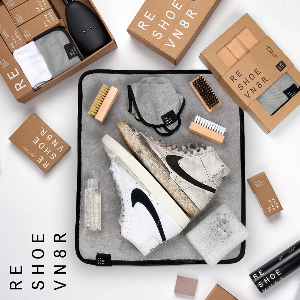 A pair of dirty most sneakers, white Nike Blazers, sit kicked back next to a box of Reshoevn8r shoe cleaning supplies on a light colored floor. The cleaning supplies include a bottle of cleaning solution, three different bristle brushes, and a microfiber towel. Text on the box indicates that the kit is suitable for suede, leather, mesh, canvas, nubuck, plastic and rubber shoes.